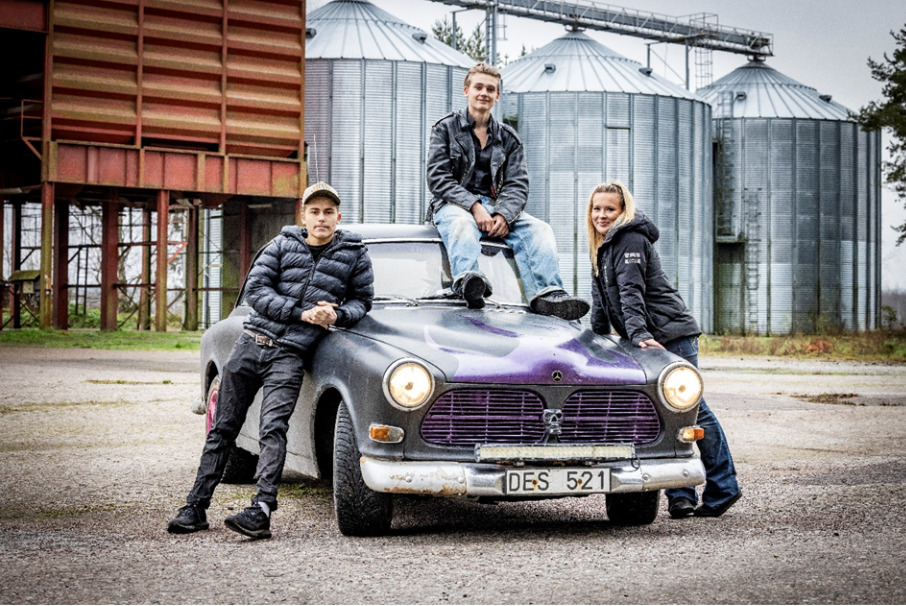 Tre ungdomar runt en gammal bil, en sitter på taket de andra två står på varsin sida